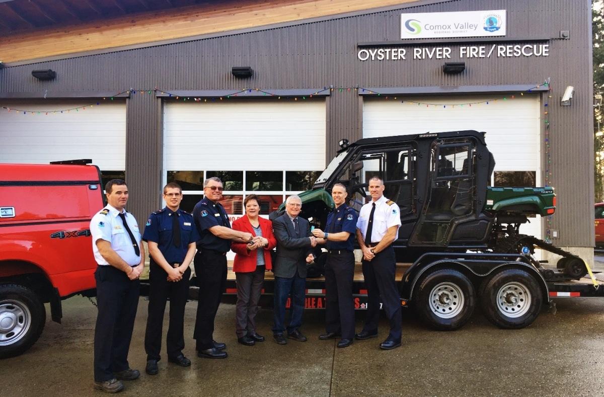 Firefighters stand with a utility vehicle on a trailer, in front of a fire station.