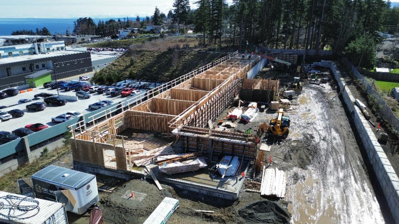 Construction site with wooden framework, machinery, and parked cars in the background.
