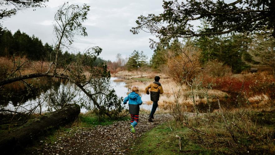 Two people walk along a riverside trail surrounded by autumn trees.