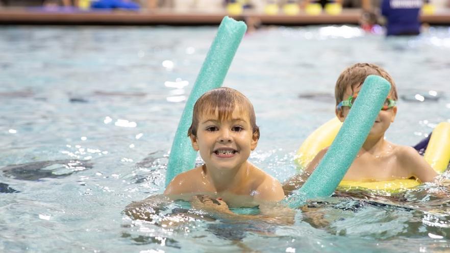 Child smiling in pool using a blue pool noodle, another child in background.
