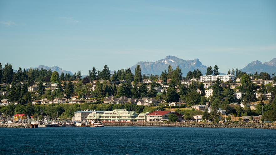 Coastal town with green hills and mountains in the background under a clear blue sky.