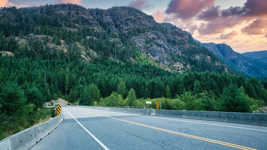 Scenic road winding through lush greenery with mountains and a colorful sunset.