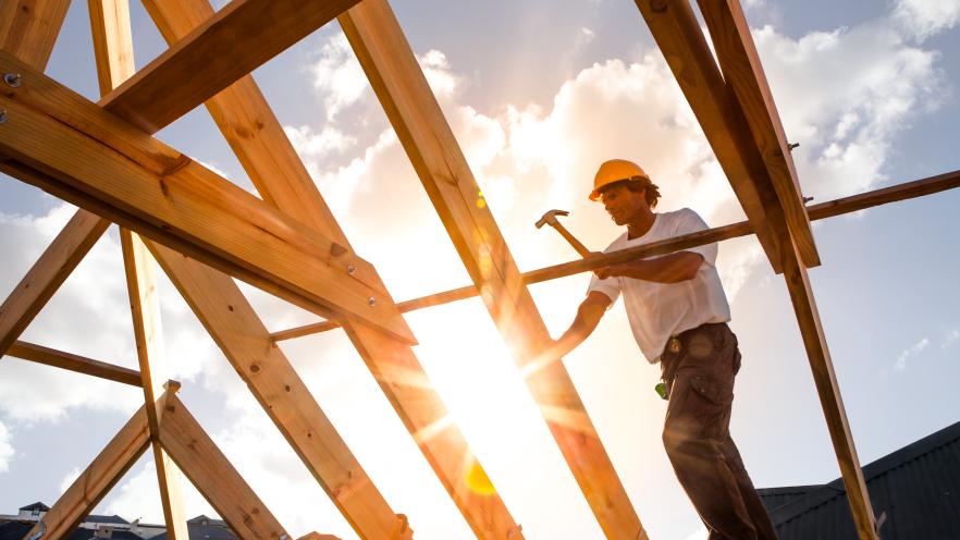 A construction worker hammers wood beams against a sunset backdrop, creating a silhouette.