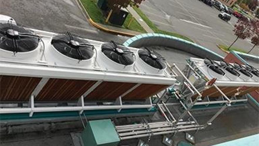 Industrial cooling units with fans atop, positioned along a rooftop, surrounded by a parking lot.