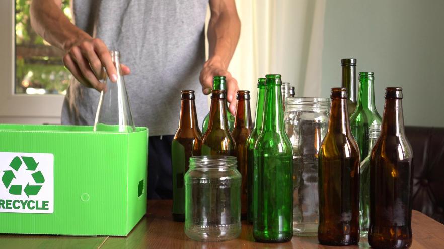 A person placing a glass bottle into a green recycling bin surrounded by various bottles on a table.