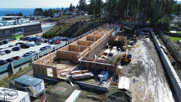 Construction site with wooden framework, machinery, and parked cars in the background.