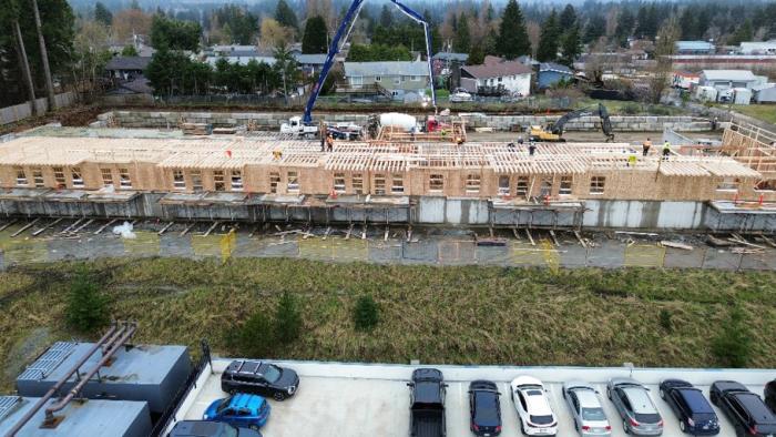 Construction site with wooden framework and machinery, surrounded by cars and greenery.