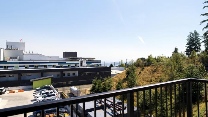 View from a balcony overlooking a forested area and a distant industrial building by the sea.