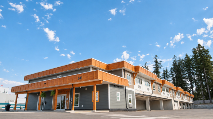 Modern multi-story building with orange accents, blue sky, and trees in the background.