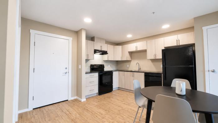 Modern kitchen with black appliances, light cabinetry, and a dining table. Natural light from windows.