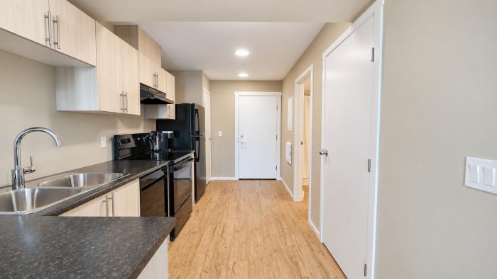 Modern kitchen with black appliances, light wood cabinets, and a spacious corridor leading to a door.