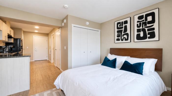 Cozy bedroom with a white bed, blue pillows, and abstract wall art, connected to a kitchen.