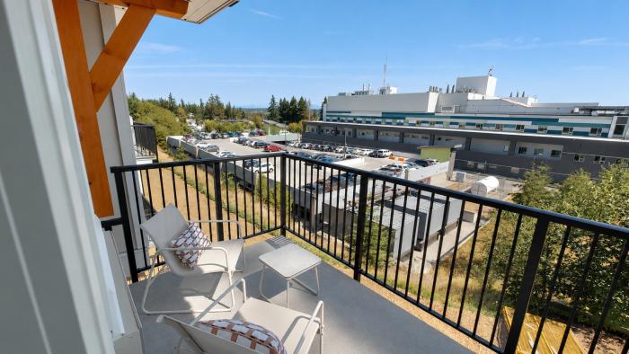Balcony view with two chairs and a table, overlooking a building and trees under a blue sky.