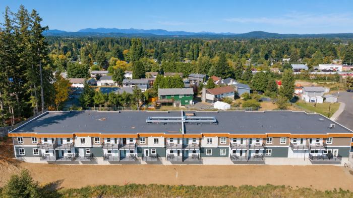 Aerial view of a modern residential building surrounded by trees and houses, under a clear blue sky.