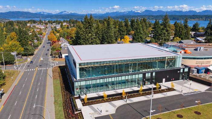 Aerial view of a modern building near a road, surrounded by trees and mountains in the background.