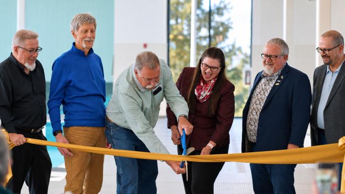 Group of six people cutting a large yellow ribbon at a celebratory event.