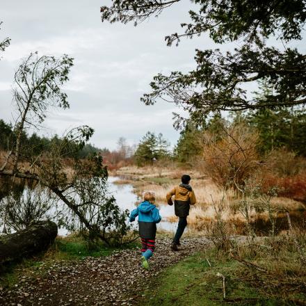 Two people walk along a riverside trail surrounded by autumn trees.