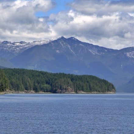 Calm water with a small boat, surrounded by lush green forests and distant mountains under a cloudy sky.