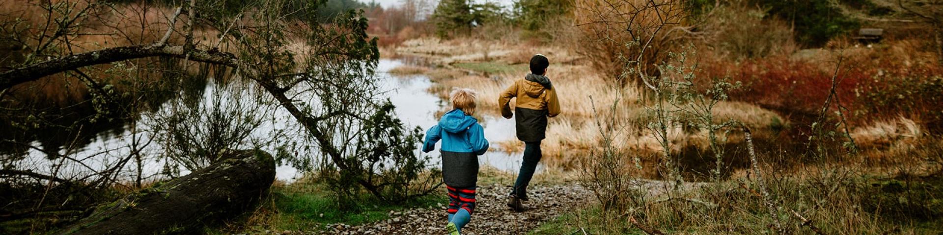 Two people walk along a riverside trail surrounded by autumn trees.
