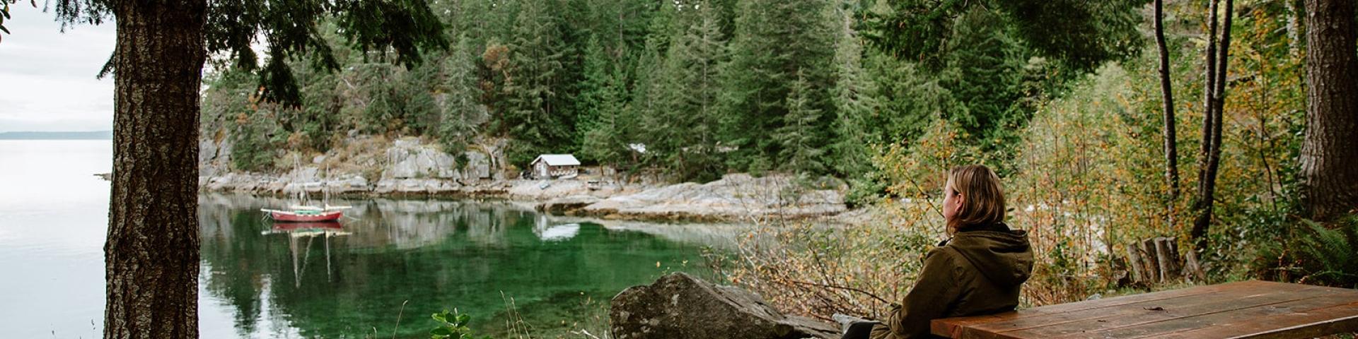 Person sitting on a picnic table by a lake, surrounded by trees.