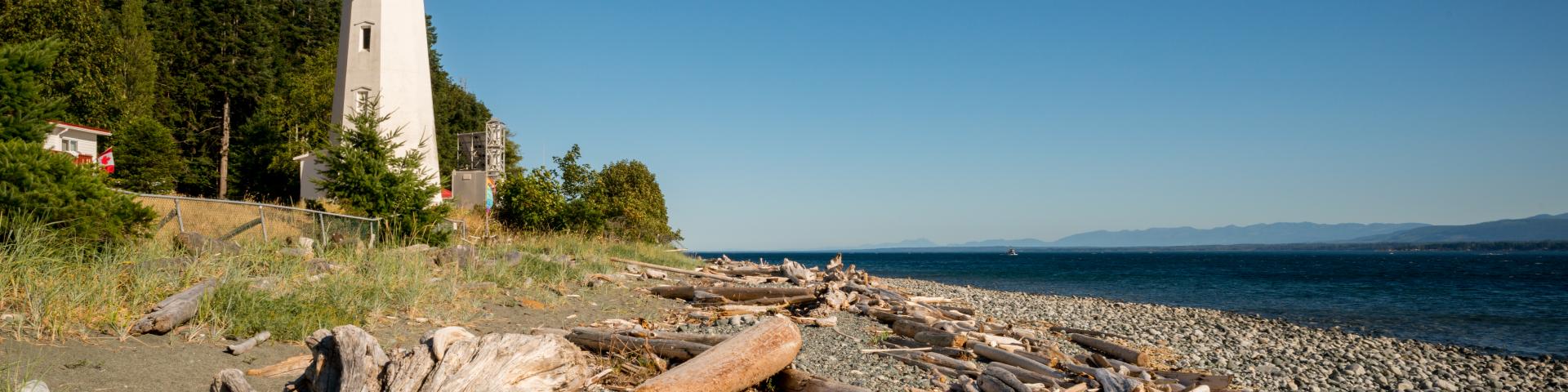 Lighthouse by a rocky beach with driftwood under a clear blue sky.