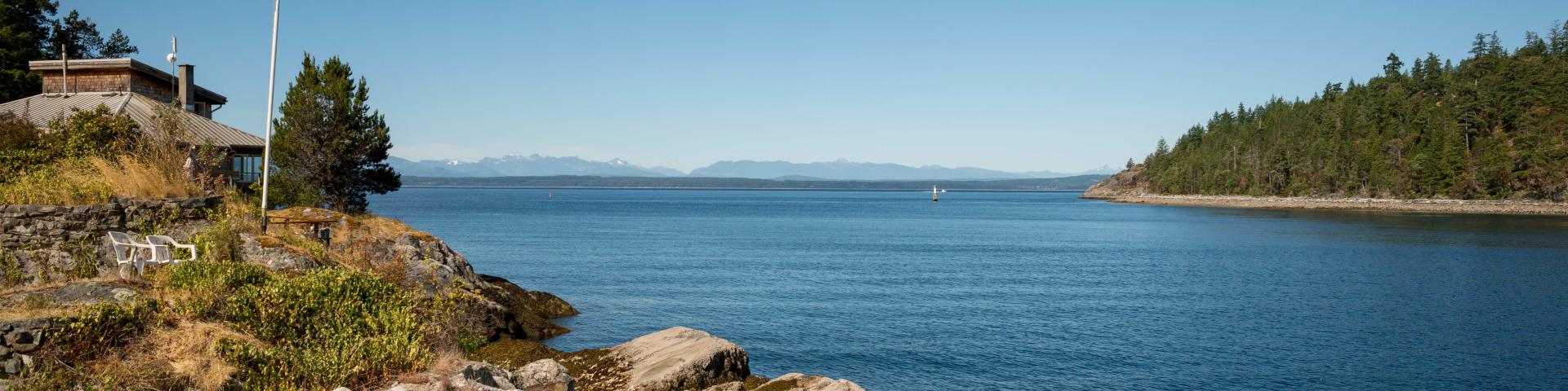 Calm blue waters beside rocky shore with Canadian flag and lush green trees in the distance.