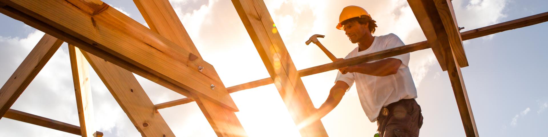 A construction worker hammers wood beams against a sunset backdrop, creating a silhouette.