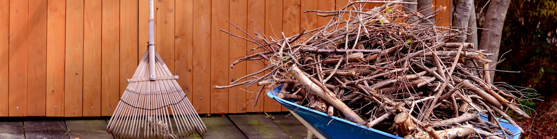 A blue wheelbarrow filled with branches next to a broom by a wooden wall.