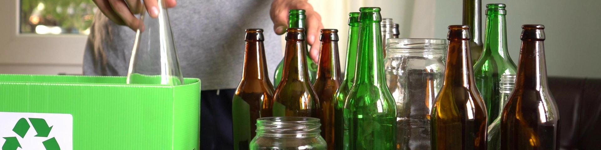A person placing a glass bottle into a green recycling bin surrounded by various bottles on a table.