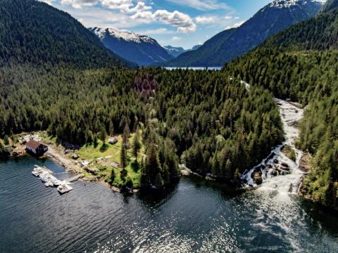 Forest and mountains surround a lakeside cabin with a waterfall nearby.