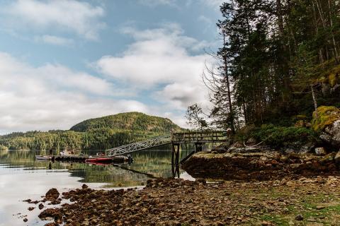 Calm lake with dock, surrounded by forest and hills under a cloudy sky.