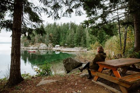 Person sitting on a picnic table by a lake, surrounded by trees.