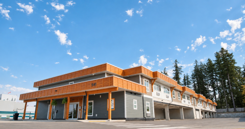 Modern multi-story building with orange accents, blue sky, and trees in the background.