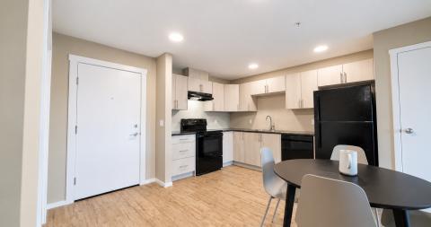 Modern kitchen with black appliances, light cabinetry, and a dining table. Natural light from windows.
