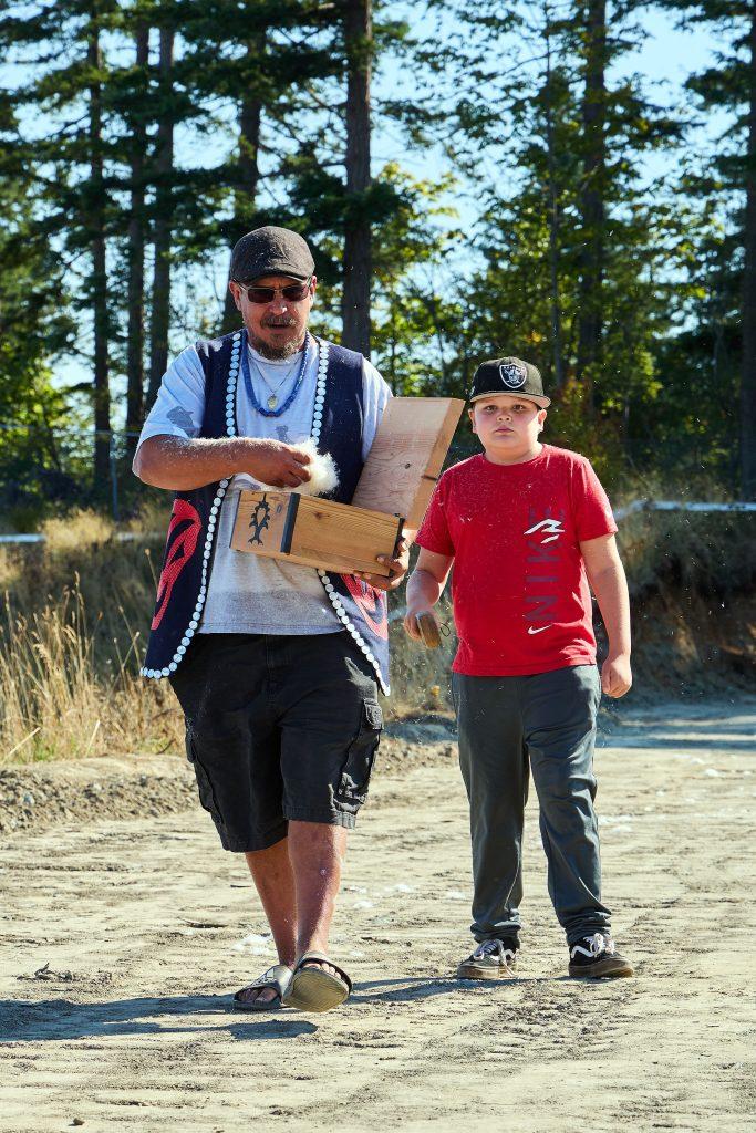 Man and child walking outdoors holding a wooden box, trees in the background.