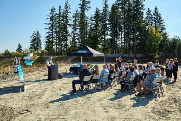 Outdoor gathering with a speaker and audience seated on a sunny day, surrounded by trees.