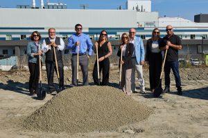 Eight people with shovels stand behind a dirt mound at a construction site.