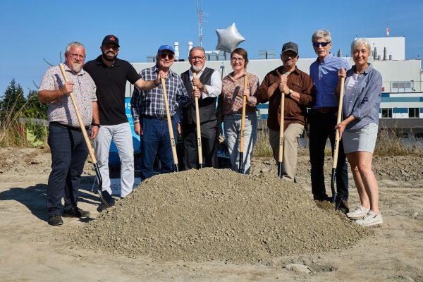 A group of people with shovels stands by a dirt mound on a sunny day.