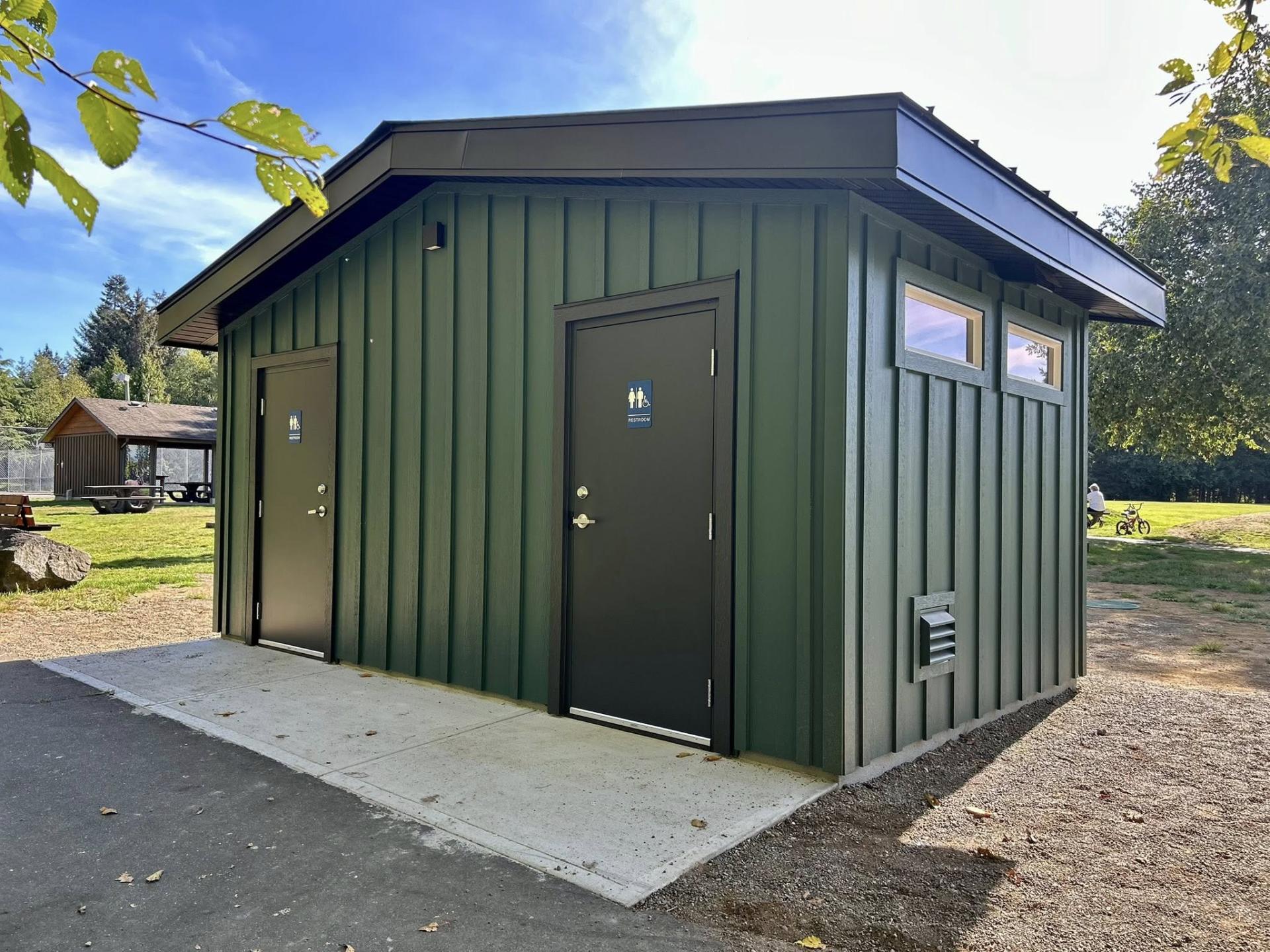 Green metal restroom building in sunny park setting.