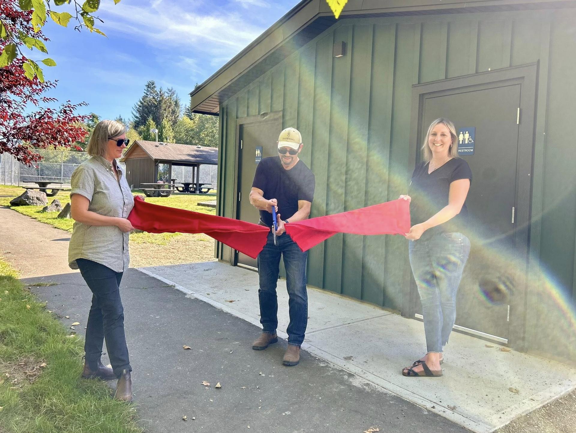 Three people cutting a red ribbon outside a bathroom building under sunny skies.