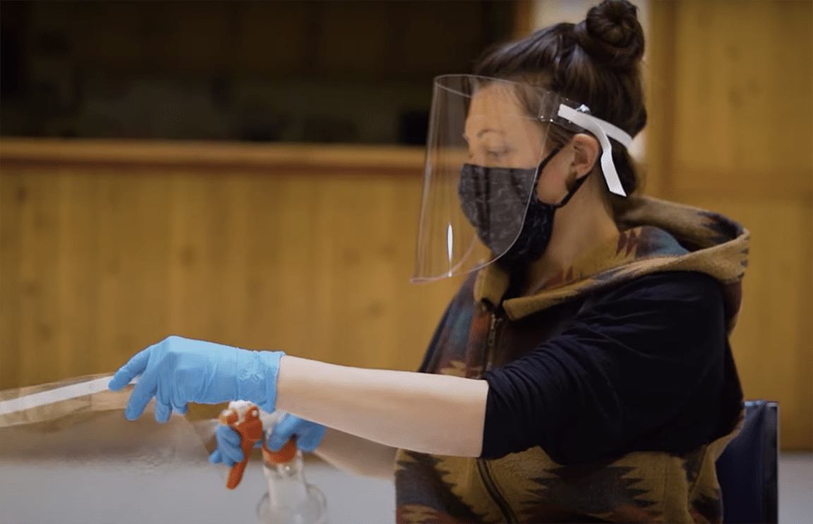 Person with face shield and mask uses a tool, wears blue gloves.