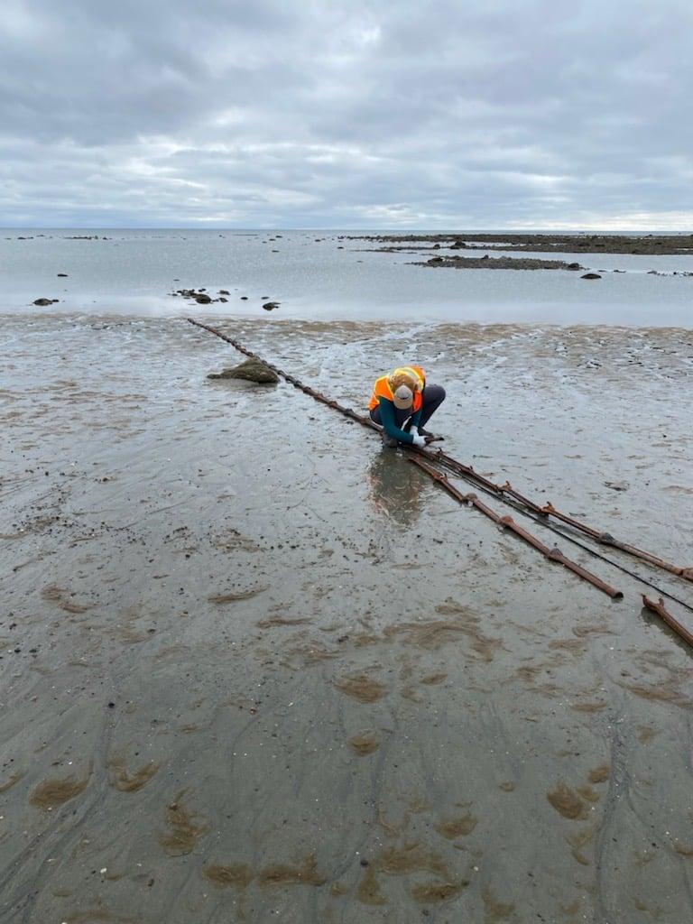 A person in an orange jacket kneels by the shore, examining driftwood at low tide.
