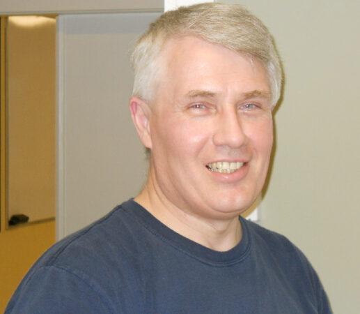 Smiling man with short gray hair in a dark shirt indoors.