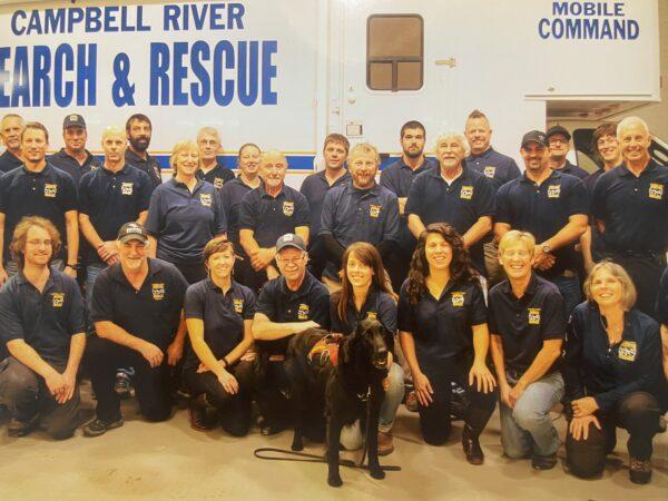 Rescue team group photo in uniform with a dog, smiling in front of command vehicle.