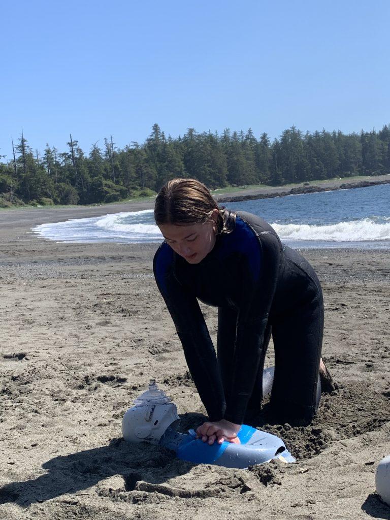 Person practicing CPR on a mannequin at a sandy beach.