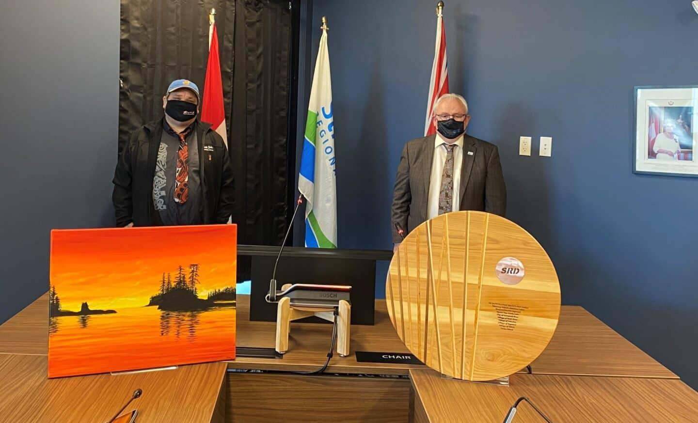 Two men in masks stand behind a table with artwork and flags.