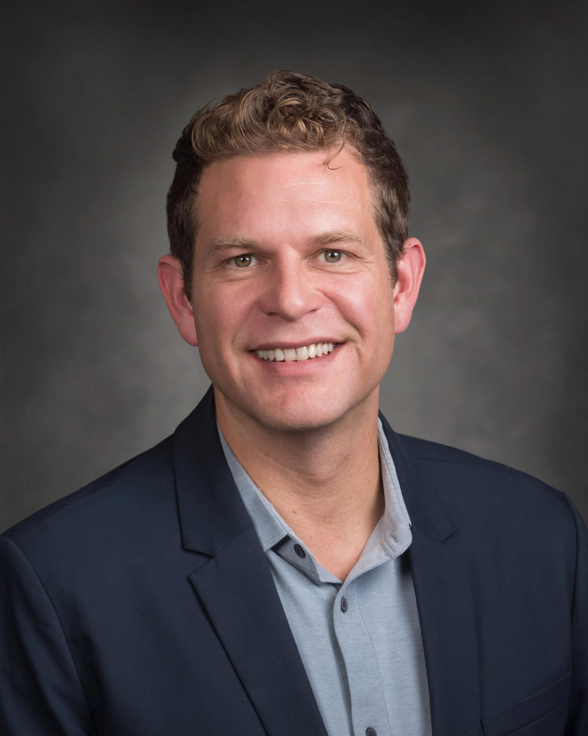 Smiling person in a suit jacket against a grey background.