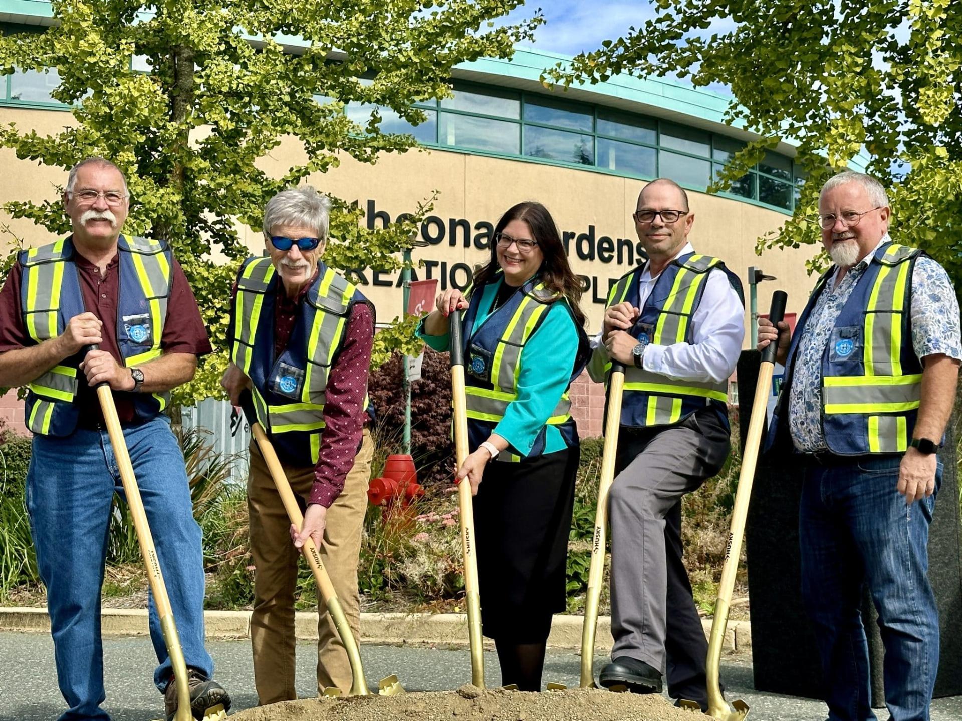 Five people with shovels wearing safety vests, standing in front of a building.