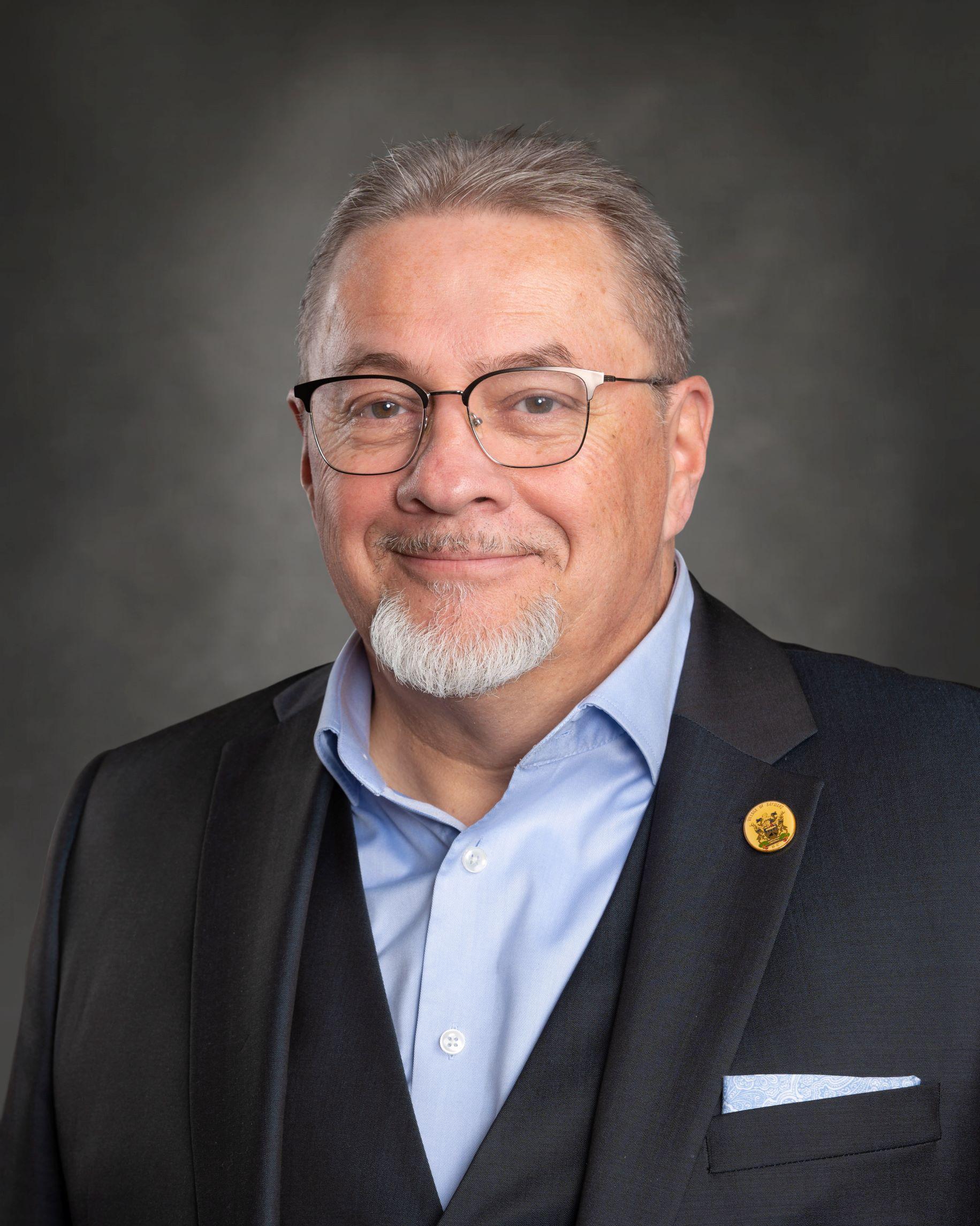 Smiling person in a suit with glasses and a goatee against a dark background.