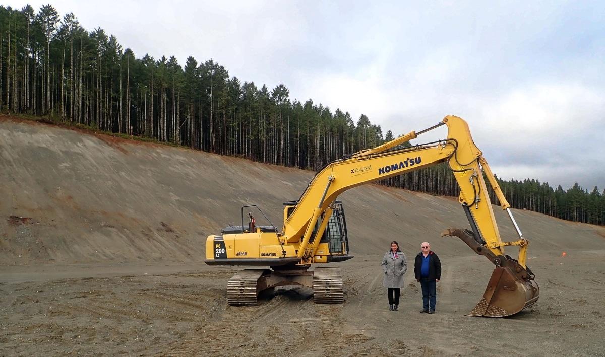 Yellow excavator on a large dirt site with two people beside it, forest in the background.
